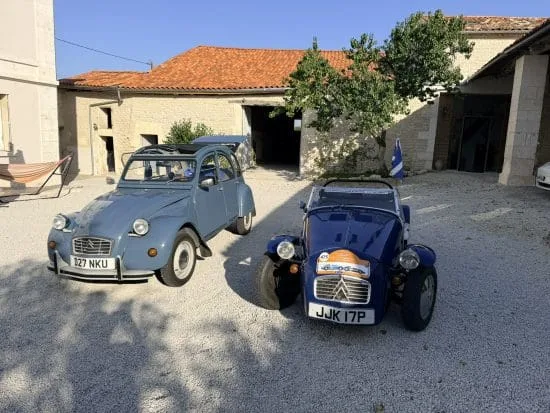 Dans un cadre enchanteur typique de la Charente, à deux pas d’Angoulême, deux voitures anciennes – une Citroën 2CV bleu clair et une élégante sportive de style Caterham ornée d’autocollants de course – attendent sur l’allée gravillonnée d’une maison d’hôtes pleine de charme. Les bâtiments en pierre aux toits de tuiles traditionnelles et le jardin verdoyant offrent une atmosphère chaleureuse, invitant à la détente lors d’un séjour authentique en Charente près d’Angoulême.