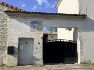 Des murs en pierre soigneusement restaurés, ornés d’une ancienne enseigne « Chocolat » légèrement effacée, surmontant une élégante porte grise et un portail en fer forgé noir, le tout adossé à de charmantes bâtisses historiques typiques de la Charente. L’ensemble évoque l’atmosphère chaleureuse d’une maison d’hôtes accueillante au cœur d’Angoulême, sous un ciel partiellement nuageux rappelant les paysages authentiques du Sud-Ouest.