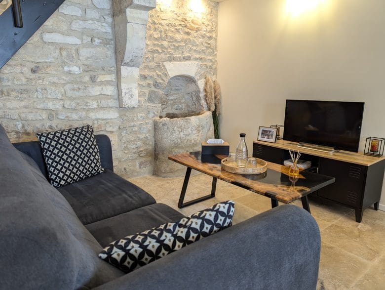 Salon chaleureux en Charente, mur en pierre, canapé gris douillet, café en bois, décoration d’Angoulême baignée d’une douce lumière accueillante.