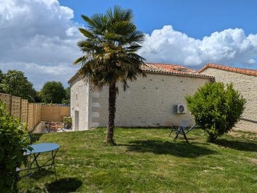 Charmante cour ensoleillée à Angoulême, en Charente, avec une pelouse verte, une table bleue, une maison en pierre typique et une ambiance chaleureuse.