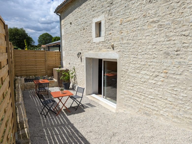 Terrasse chaleureuse en Charente : gravier, deux tables orange, chaises noires, plantes et maison typique près d’Angoulême, ciel nuageux.