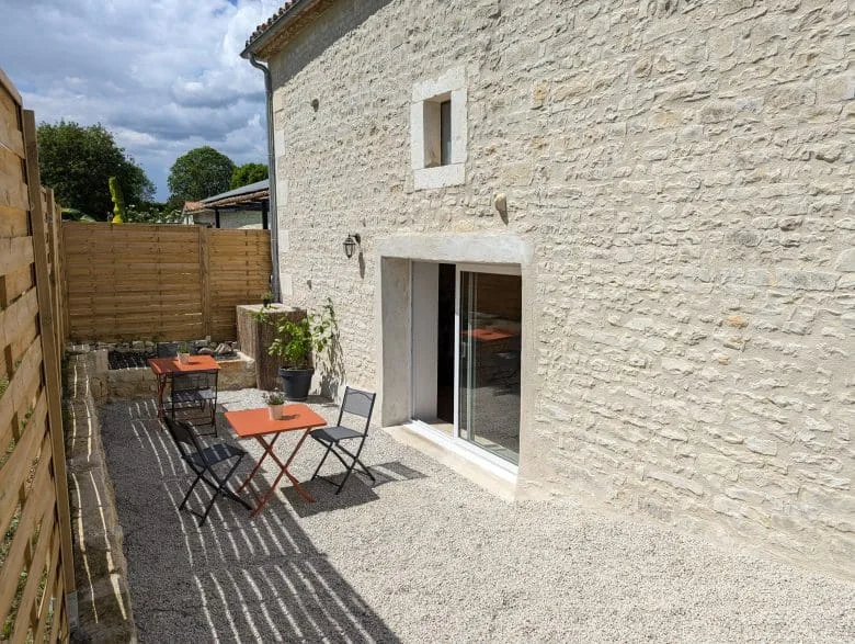 Terrasse chaleureuse en Charente : gravier, deux tables orange, chaises noires, plantes et maison typique près d’Angoulême, ciel nuageux.