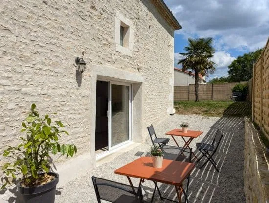 Charmante maison en pierres de Charente, patio chaleureux près d’Angoulême, tables colorées, citronnier et palmier sous un ciel lumineux.