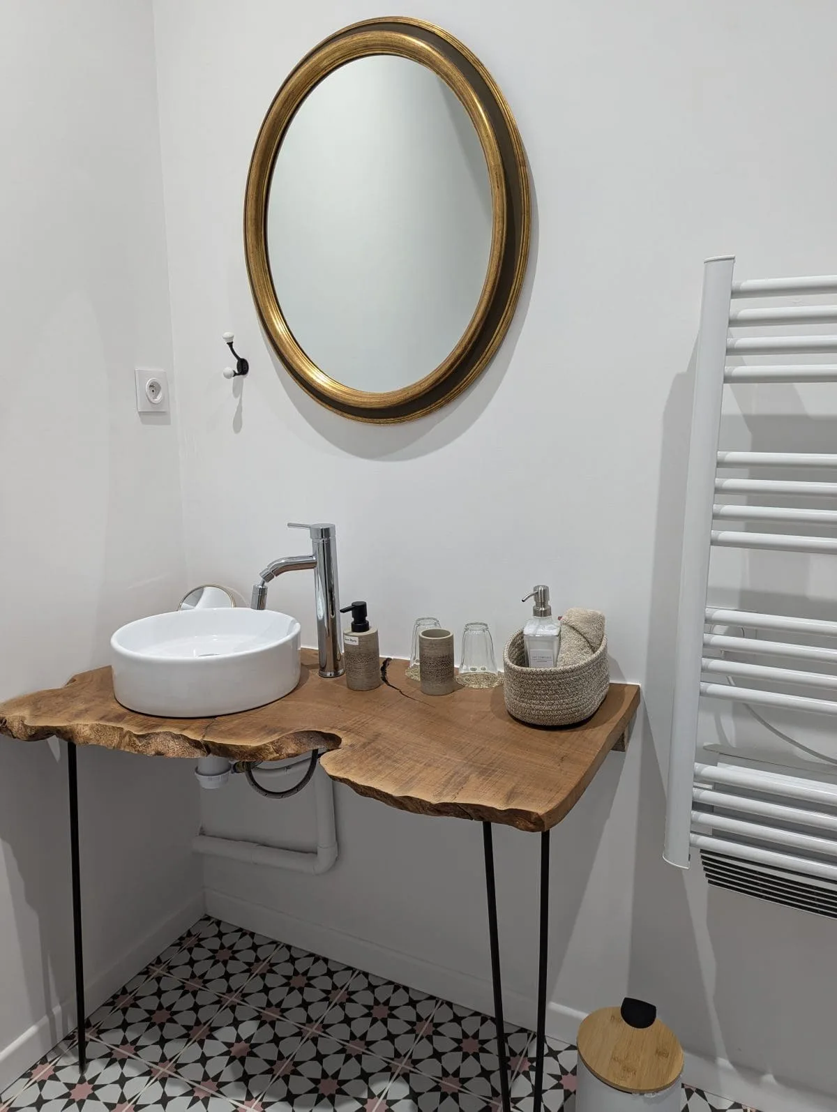 Dans une demeure typique de la Charente à Angoulême, une salle de bains chaleureuse : miroir doré, vasque en bois, panier en osier, carrelage décoratif.