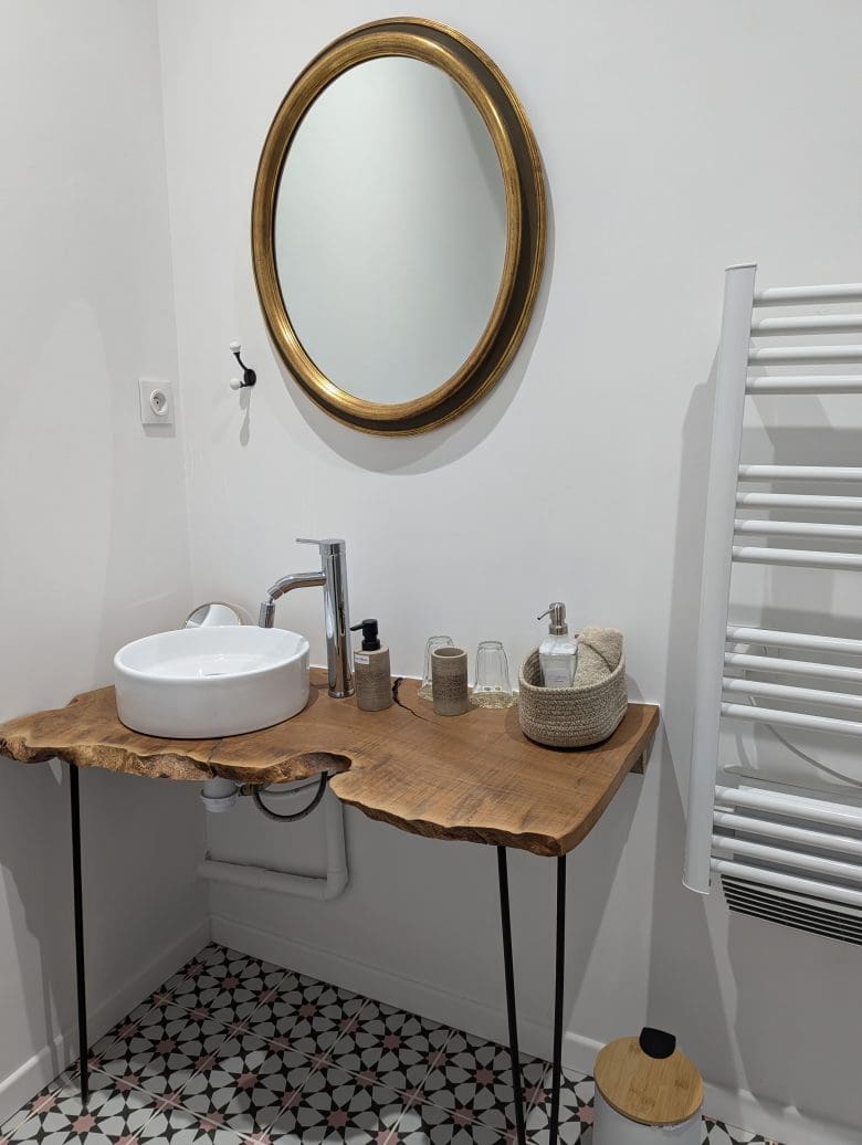 Dans une demeure typique de la Charente à Angoulême, une salle de bains chaleureuse : miroir doré, vasque en bois, panier en osier, carrelage décoratif.