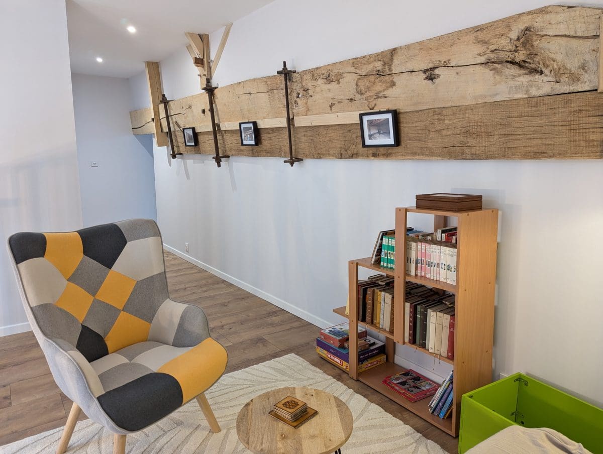 Salon chaleureux d'une maison d'hôtes en Charente, avec un fauteuil en patchwork, une table ronde, une étagère à livres et une poutre rustique à Angoulême.