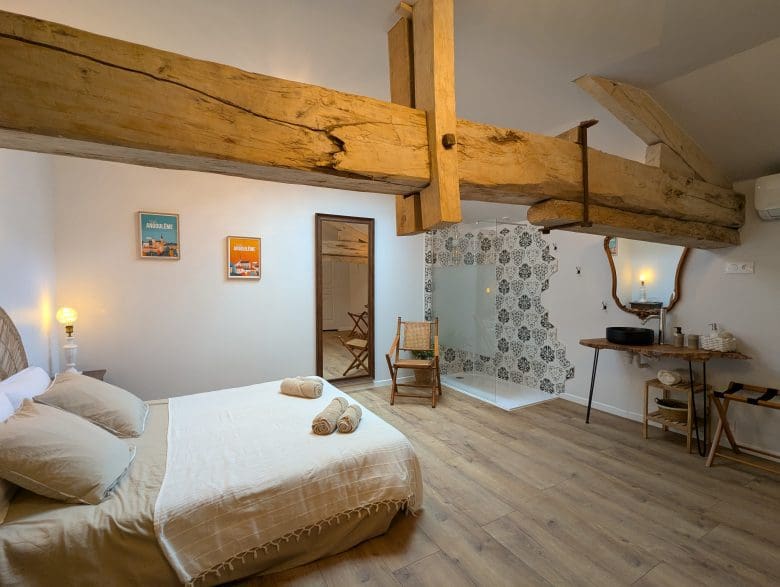 Chambre d’hôtes chaleureuse en Charente, avec poutres apparentes, lit double, fauteuil, miroir et salle de bains élégante, près d’Angoulême.