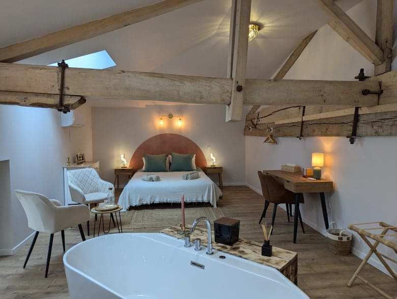 Chambre chaleureuse dans le style d’une maison d’hôtes en Charente : poutres apparentes, lit double, baignoire, coin salon et bureau près d’Angoulême.