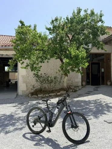 Un VTT noir garé sur une allée de gravier devant une authentique bâtisse charentaise aux murs en pierres apparentes, typique de la région d’Angoulême. Un arbre aux larges feuilles offre une ombre rafraîchissante près de cette charmante maison d’hôtes au toit de tuiles traditionnelles et à la porte en bois, offrant un cadre accueillant et reposant pour découvrir la Charente.