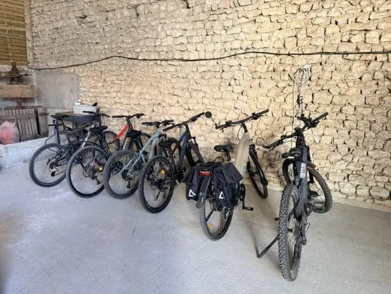 Dans notre chaleureuse maison d’hôtes, six vélos de styles et de tailles variés sont soigneusement alignés à l’intérieur contre un authentique mur en pierre de Charente, qui rappelle le charme du patrimoine angoumoisin. Parfaits pour explorer Angoulême et ses environs pittoresques à vélo, certains vélos sont équipés de sacoches pratiques pour vos balades le long des vallées charentaises. Le sol en béton apporte une touche industrielle élégante, tandis qu’une structure métallique sur la gauche complète ce garage dédié aux cyclistes désireux de découvrir la région sous son meilleur jour.