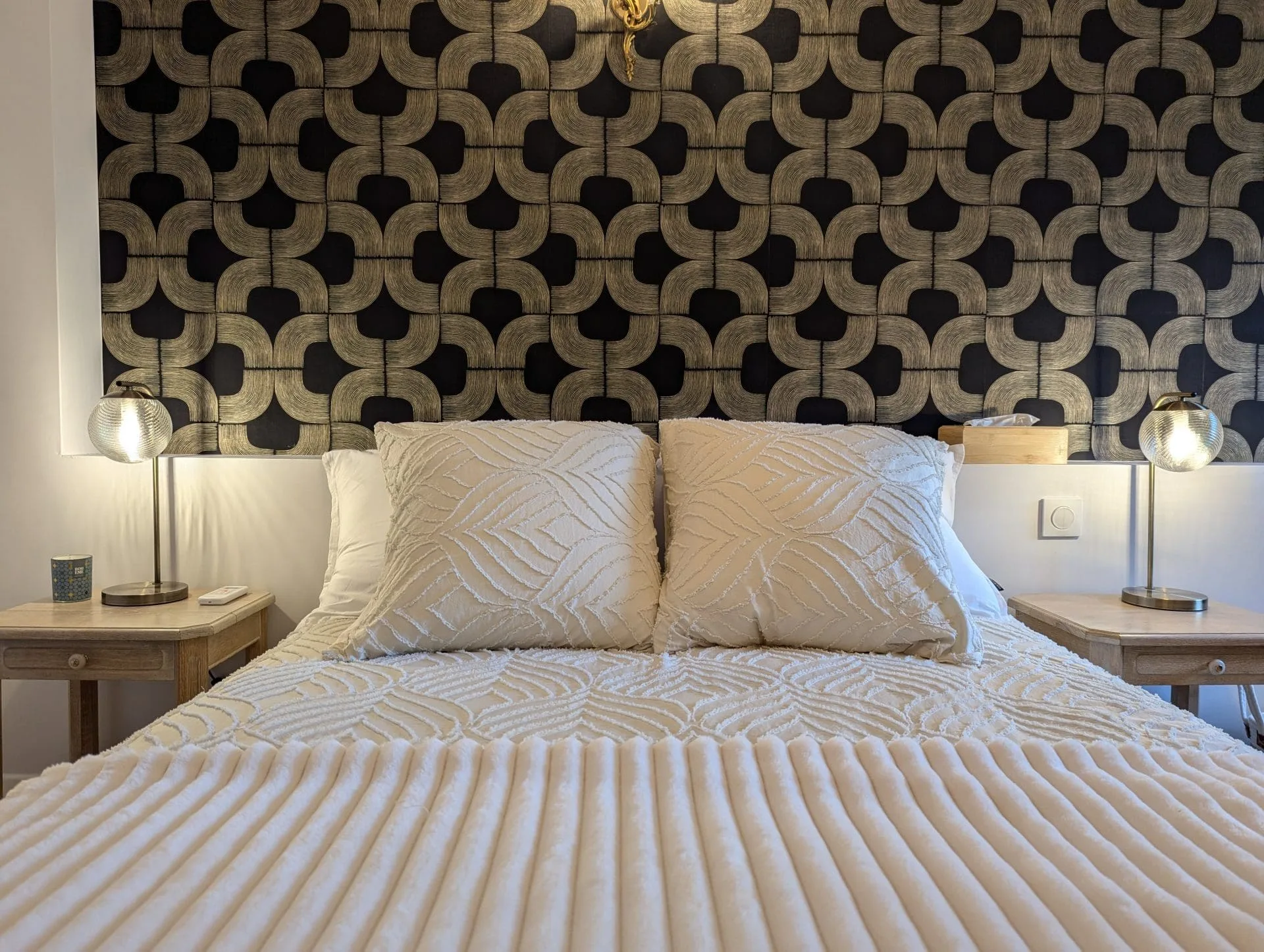 Dans une chambre chaleureuse inspirée des chambres d’hôtes de Charente, près d’Angoulême, un lit soigneusement préparé vous invite au repos avec deux oreillers blancs texturés et une élégante couette assortie. De part et d’autre, de charmantes tables de chevet en bois agrémentées de lampes aux lumières douces rappellent l’accueil authentique du Sud-Ouest. Le papier peint sombre à motifs donne du caractère et met en valeur l’ambiance raffinée qui fait le charme des hébergements typiques autour d’Angoulême.