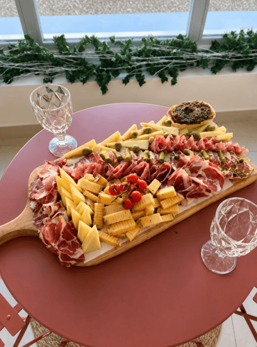Plateau de charcuterie convivial typique de la Charente, composé d’un assortiment généreux de viandes fines locales, de fromages affinés d’Angoulême, de gressins artisanaux, de tomates cerises du marché et de tapenade maison, présenté sur une table rouge chaleureuse. Deux verres en cristal vides invitent à la dégustation devant une fenêtre ornée de lierre vert, dans l’ambiance authentique et accueillante d’une maison d’hôtes en Charente près d’Angoulême, idéale pour savourer les spécialités gastronomiques régionales.